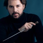 Conductor Simon Rivard is looking at the camera and holding a conducting baton. He has brown hair and a beard and is wearing a black turtleneck