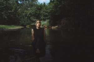 Sarah Kirkland Snider is wearing a black dress and standing in a creek. She has long blonde hair.