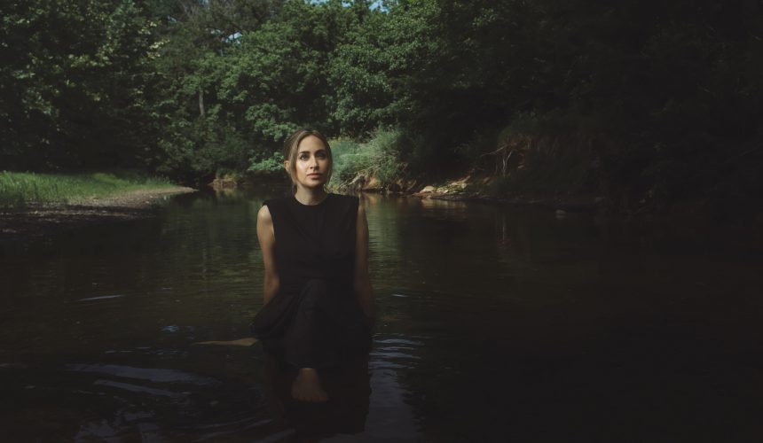 Sarah Kirkland Snider is wearing a black dress and standing in a creek. She has long blonde hair.